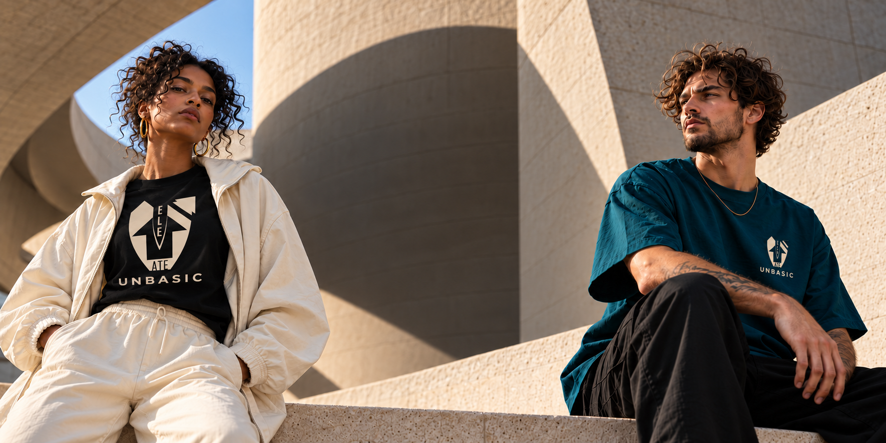 Two models among sunlit stone architecture: a woman with a centered ivory Elevate Unbasic graphic on a black tee and a man with a small ivory chest logo on a deep teal tee
