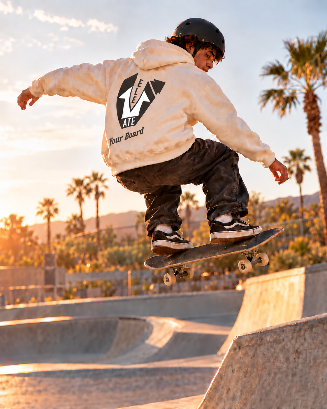 A teenage skateboarder in mid-air above a skatepark ramp, wearing an ivory hoodie with the charcoal and white Elevate design and small Your Board lettering following the hoodie’s folds