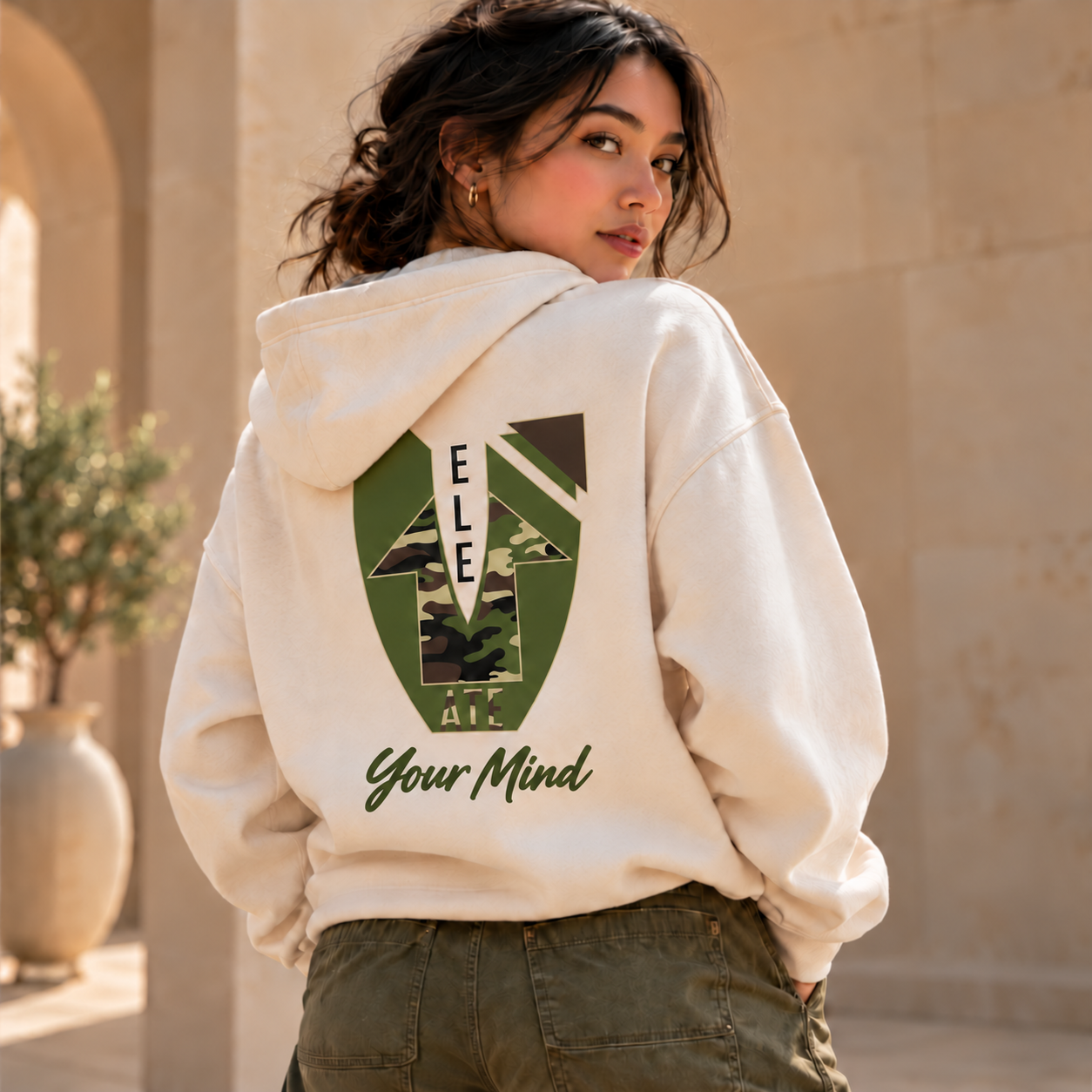 A woman wearing the cream Elevate Your Mind hoodie, with the custom camouflage V and bold olive script